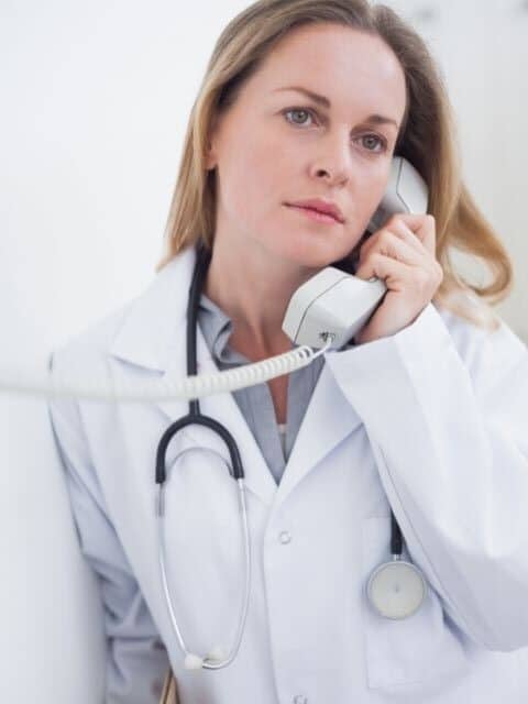 Female doctor in a white lab coat with a stethescope around her neck holding a white, corded phone up to her ear while she looks to the right of the camera.