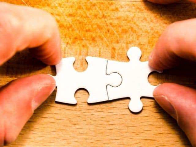 Close up view of two thumbs and two index fingers fitting two solid white puzzle pieces together.