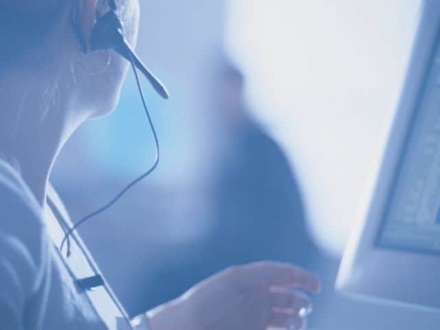Close up of the right side of a woman's head looking away from the camera while wearing a black phone earpiece around her right ear. The edge of a computer monitor is on the right side of the image and the background is blurred.