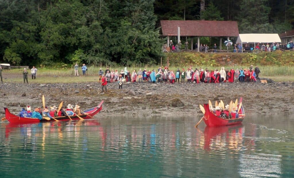 Two red Tlingit Canoes arrive in Bartlett Cove Alaska with a group of onlookers on the shore watching them.
