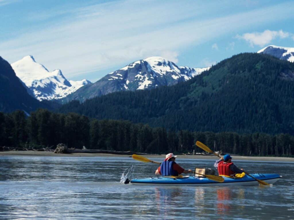 Two men kayaking in Alaska on calm waters on a sunny day. Snowcapped mountains and a pine forest are in the background.