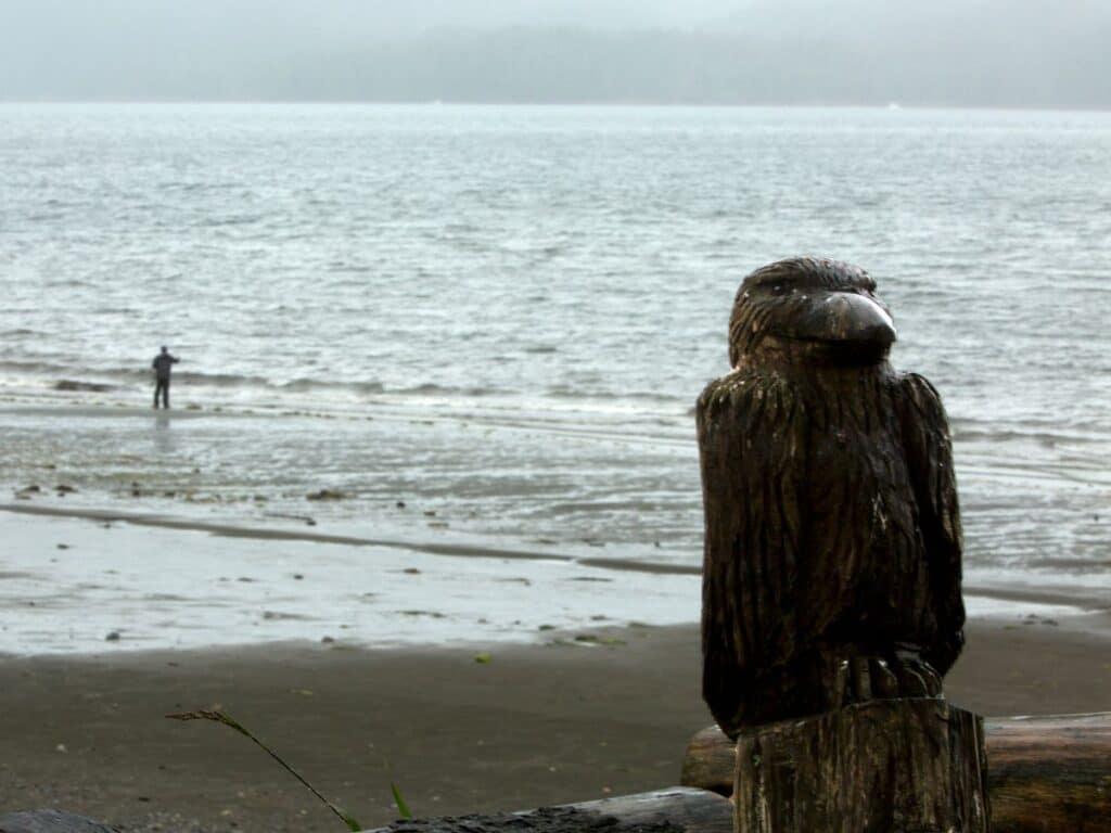 Tlingit fisherman off in the distance on a beach in Alaska, with a close up view of a wooden carved raven totem.