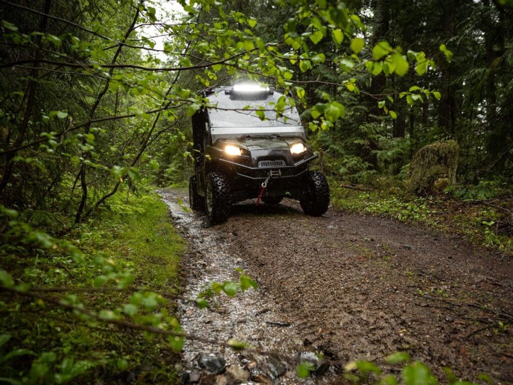 UTV driving down a dirt road with its headlights on in a green deciduous forest.