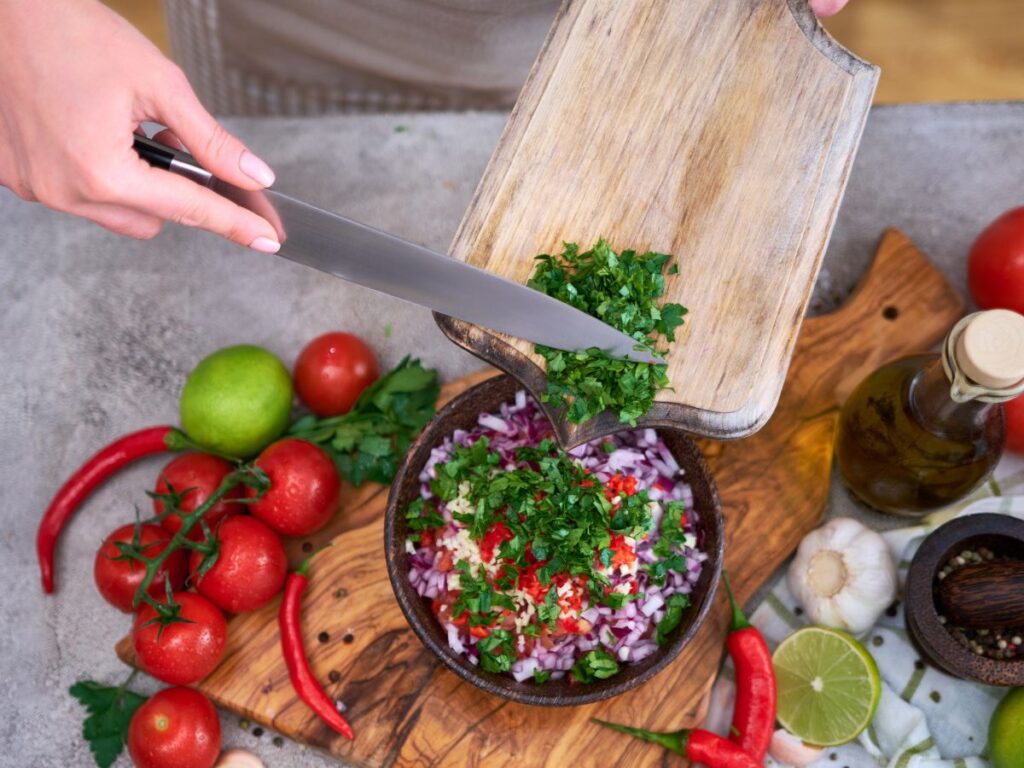 hand scraping chopped cilantro into pico de gallo from a wooden cutting board
