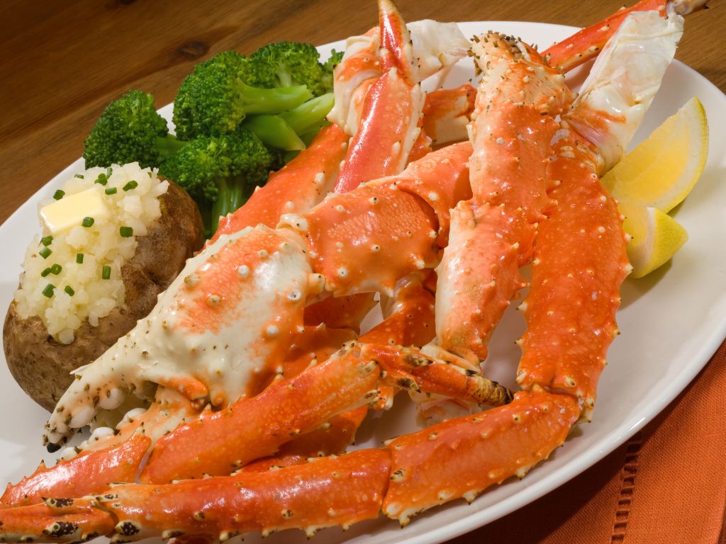 King crab legs on a plate with a baked potato, broccoli, and a lemon wedge.