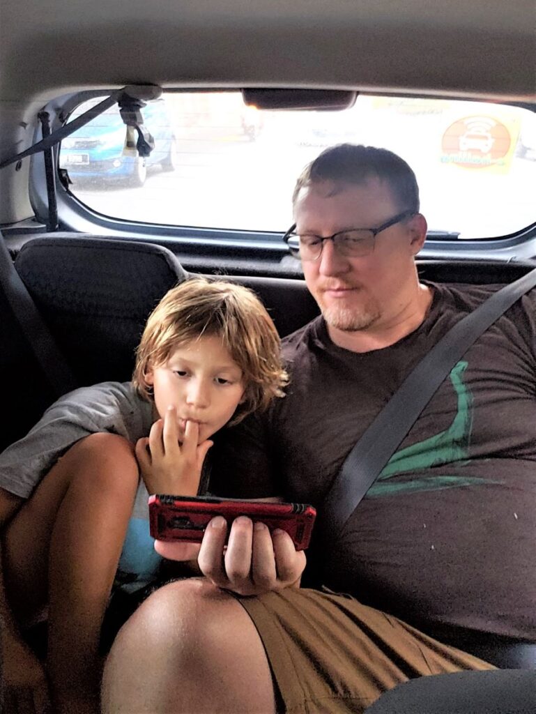 David and Little Man sitting in the backseat of an Uber while watching a video on David's phone, which David is holding.