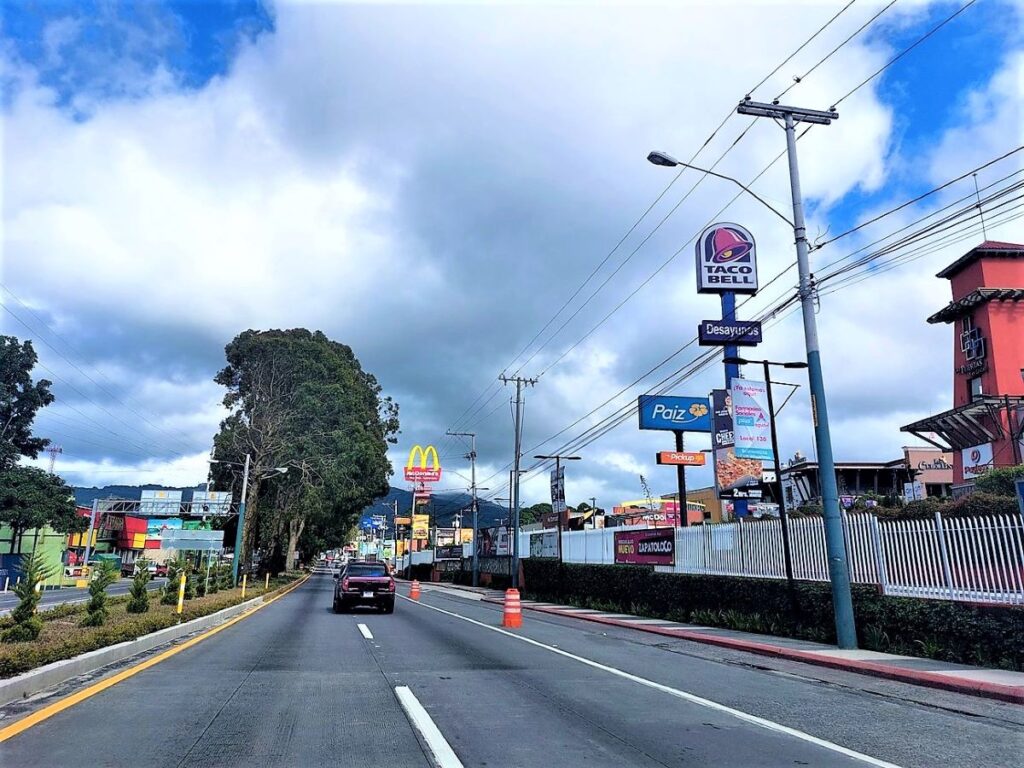 Street in Guatemala City. A pickup truck is driving the same direction the camera is facing. Various fast food restaurants, including McDonalds and Taco Bell, are on the right side.