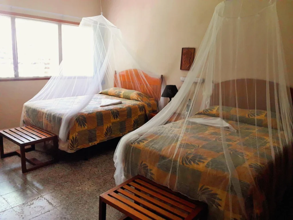 Guest room with two double beds at Tikal Inn in Guatemala. There are mosquito nets over the beds and short tables at the end of each bed with a single nightstand with a lamp in between the beds.