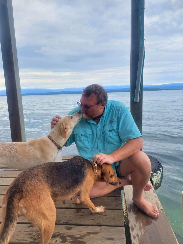 David sitting on a dock with two dogs for a house sit in Bocas del Toro, Panama. He is petting the head of a mid-sized brown and black dog and getting a kiss on the face by a white and light tan dog.
