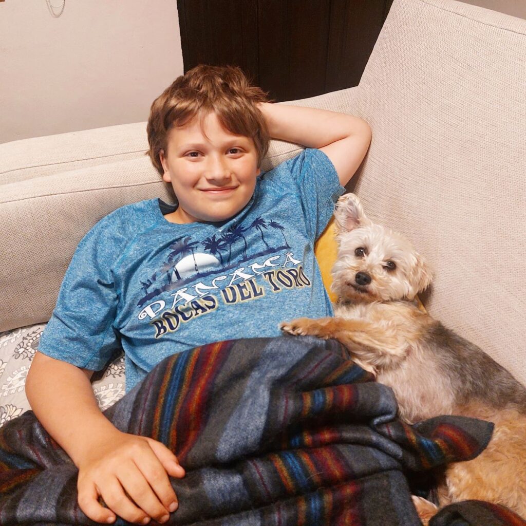 Little Man sitting on a couch under a blanket up to his waist, with a small dog laying contentedly next to him, at a house sit in Ecuador.