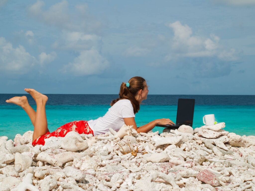 Woman laying on the beach while typing on her laptop overlooking the ocean.